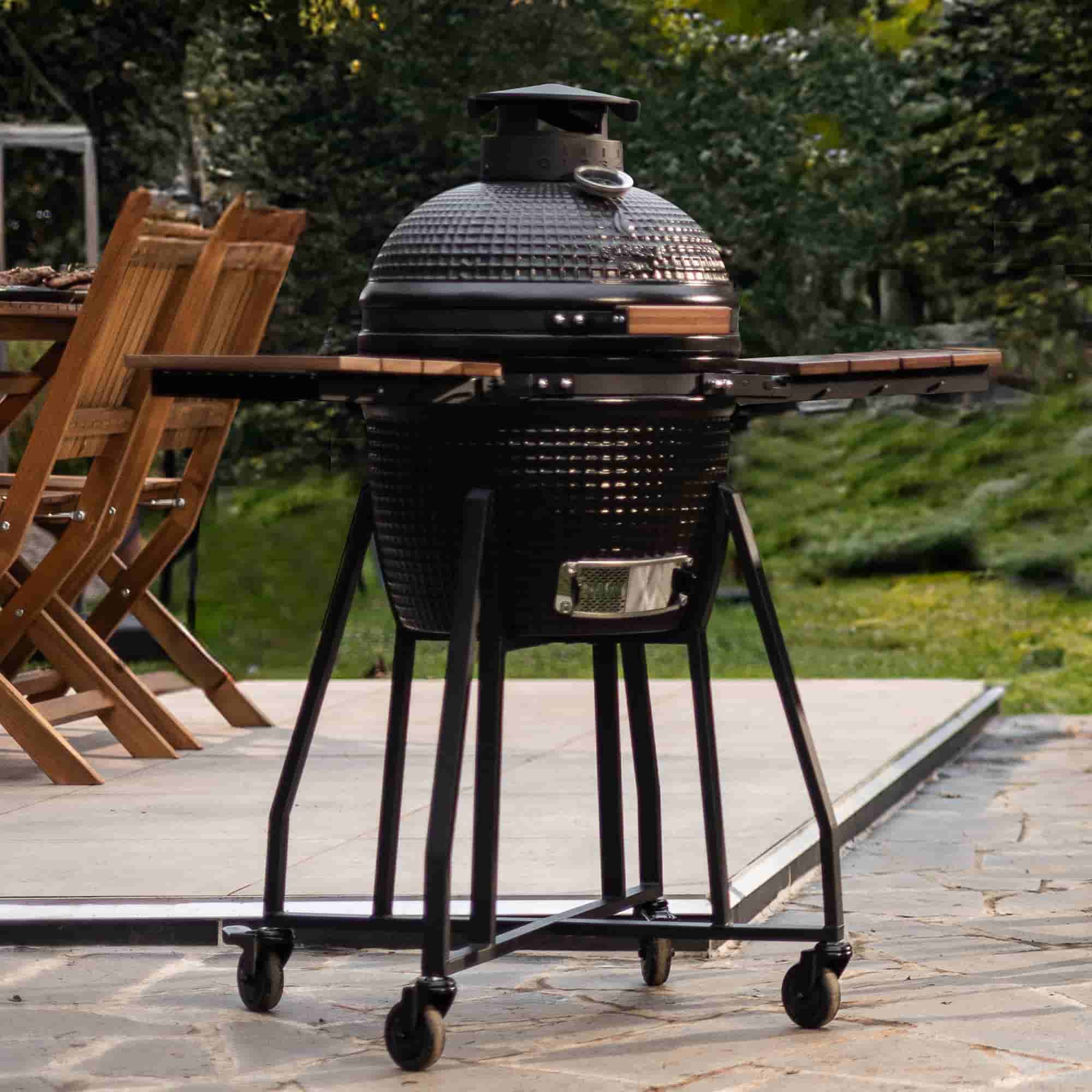 Barbecue kamado noir sur chariot à roulettes avec tablettes latérales en bois, installé sur une terrasse extérieure près d’une table en bois.