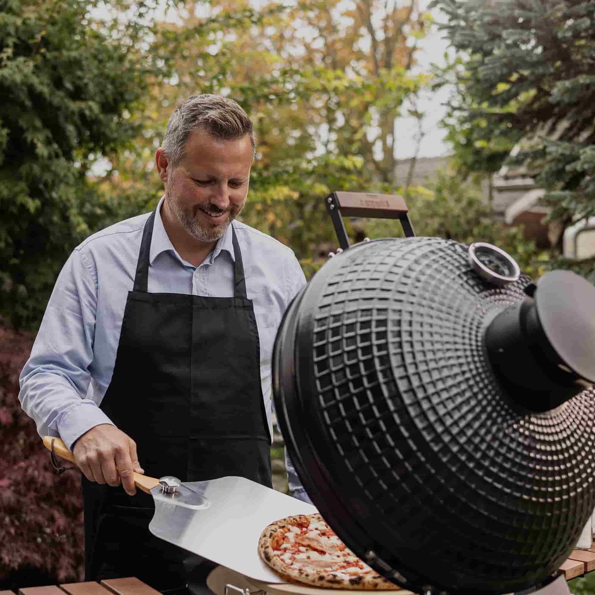 Personne portant un tablier noir sortant une pizza d’un barbecue kamado à l’aide d’une pelle à pizza, en extérieur dans un jardin.
