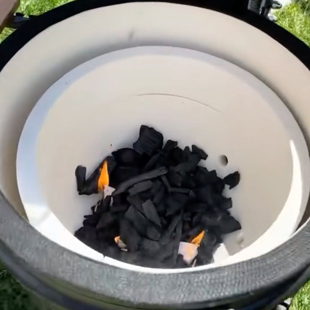 Vue intérieure d’un barbecue Kamado Forest Grill avec le foyer en céramique blanche, contenant du charbon de bois noir en train de s’enflammer légèrement, illustrant la phase d’allumage du barbecue.
