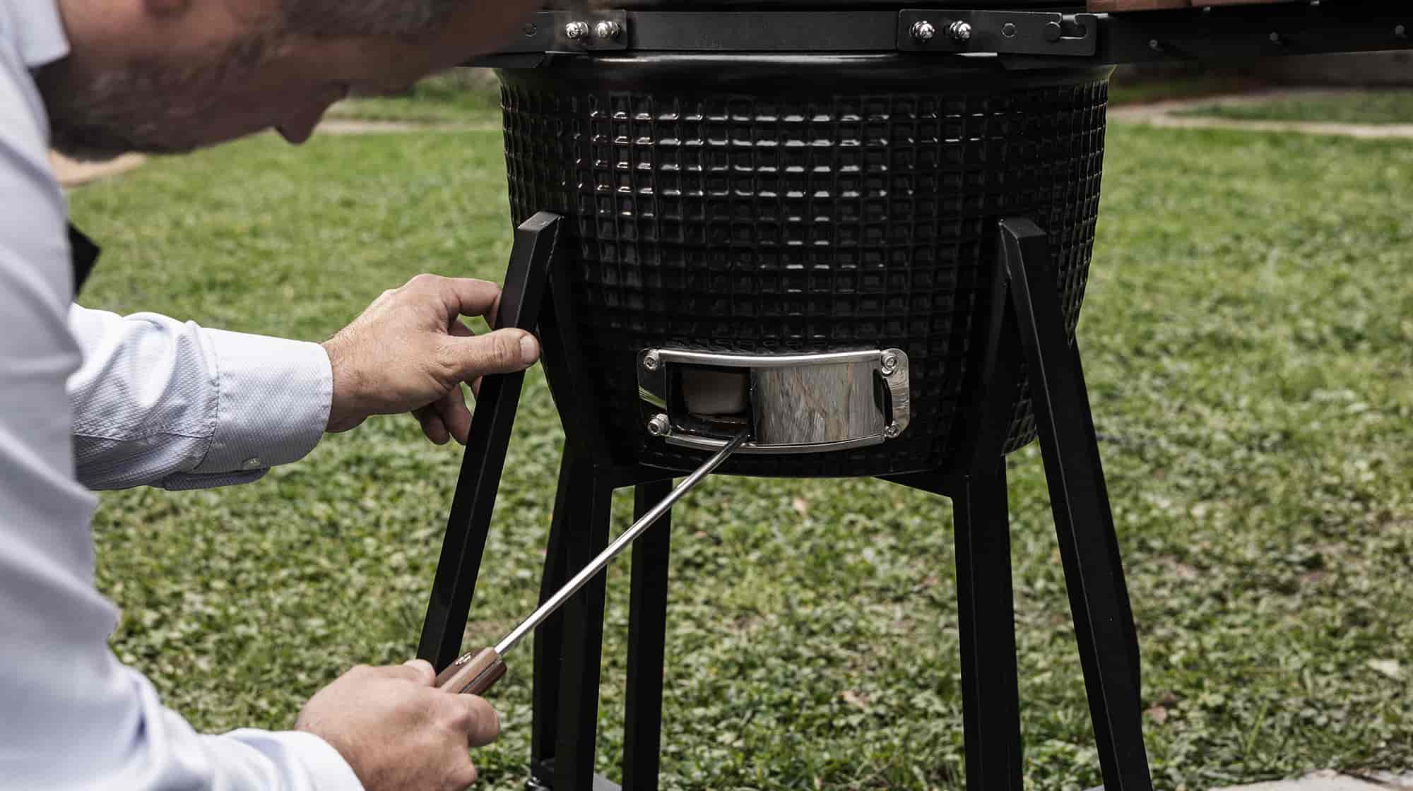 Personne nettoyant le cendrier en inox du barbecue Kamado Forest Grill à l’aide d’un tire-cendre.