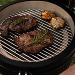 Gros plan sur la cuisson de deux steaks garnis de romarin et de têtes d’ail grillées sur la grille en inox d’un barbecue Kamado Forest Grill.