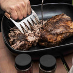 Main utilisant une griffe à viande en acier inoxydable pour effilocher un rôti juteux dans un plat noir, sur une table de barbecue avec épices en arrière-plan.