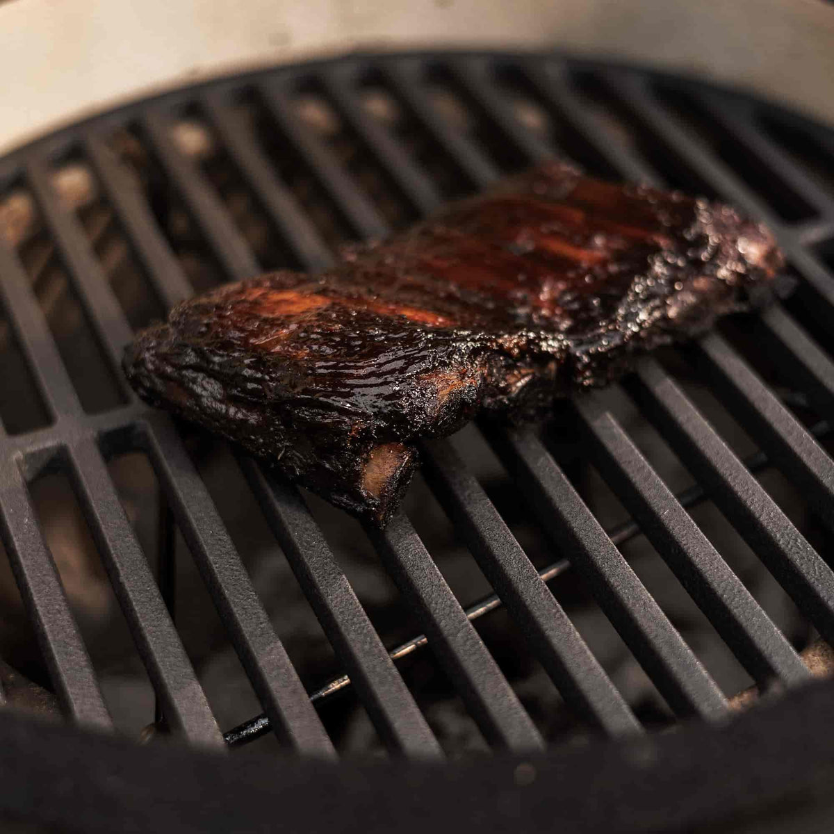 Pièce de viande grillée et caramélisée posée sur une grille de cuisson en fonte noire à l’intérieur d’un barbecue Kamado.