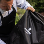 Gros plan d’une personne plaçant une housse noire sur un barbecue Forestgrill, avec le logo de la marque visible au centre du tissu.