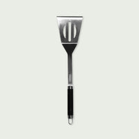 Long stainless steel spatula with ergonomic black handle and hanging hole, ideal for turning food on the barbecue or plancha.