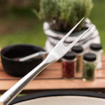 Gros plan sur une fourchette de barbecue en acier inoxydable avec manche en bois, tenue au-dessus d’un Kamado avec des épices en arrière-plan.