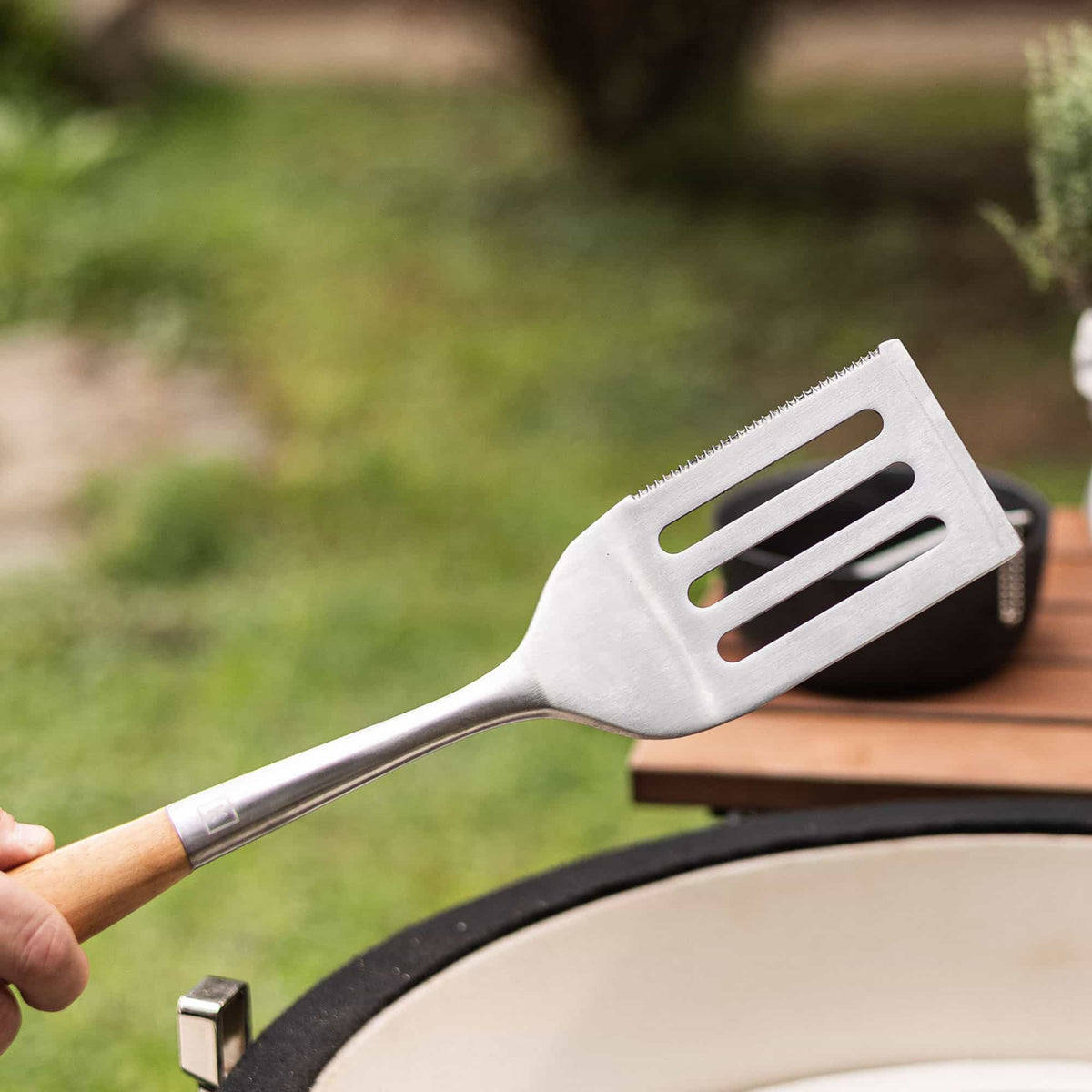 Main tenant une spatule de barbecue en acier inoxydable avec manche en bois, au-dessus d’un Kamado ouvert dans un jardin.