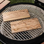 Deux planches rectangulaires en bois d’olivier posées sur la grille d’un barbecue kamado, prêtes à être utilisées pour le fumage et la cuisson des aliments.