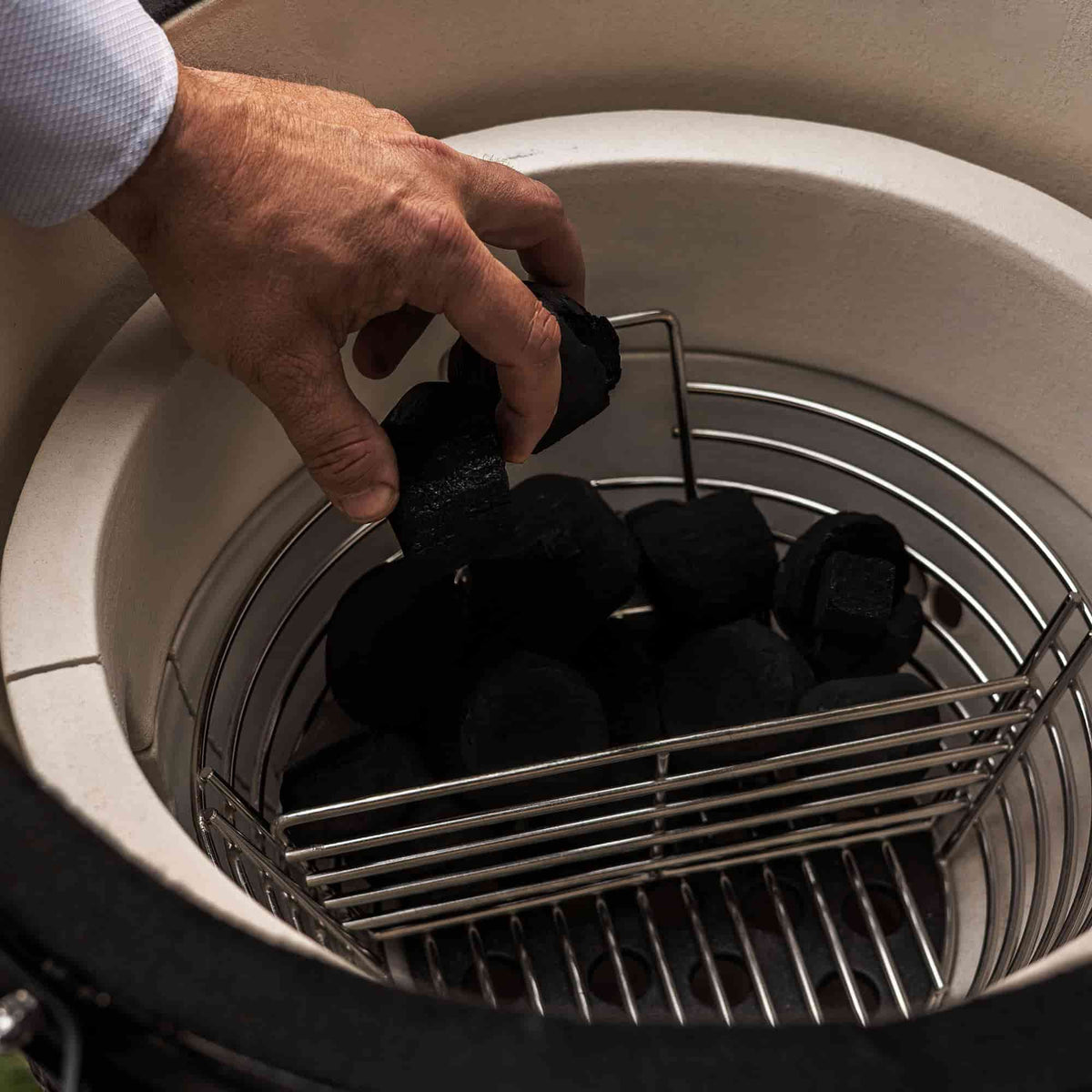 Main plaçant des morceaux de charbon dans un panier en acier inoxydable à l’intérieur d’un barbecue Kamado, avant l’allumage pour la cuisson.