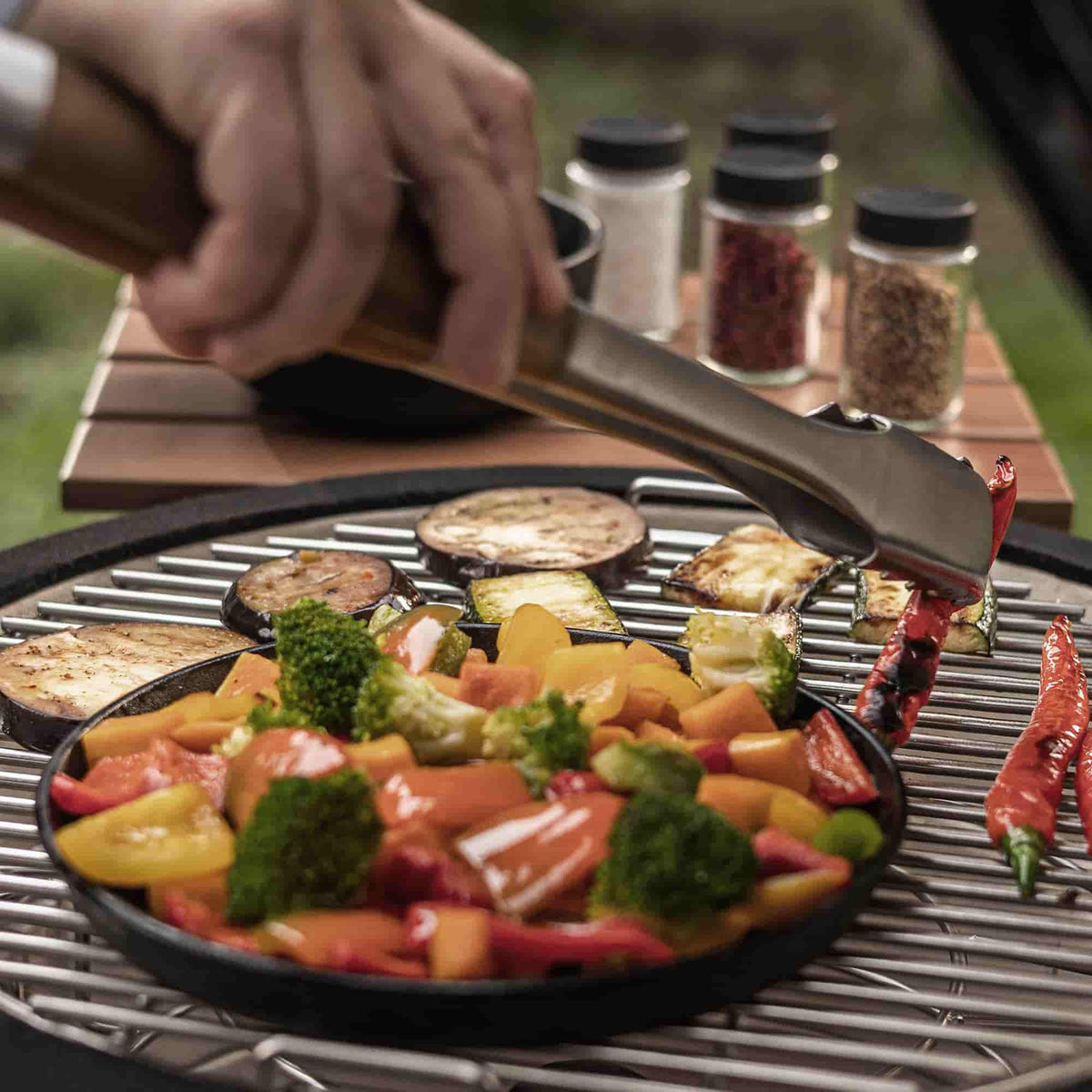Légumes colorés en train de griller sur un Kamado, disposés dans une poêle en fonte noire ; une main utilise une pince de cuisson en inox pour retourner les aliments, avec des épices visibles en arrière-plan.