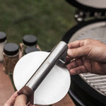 Presse à burger en acier inoxydable tenue en main devant un barbecue Kamado, avec plusieurs pots d’épices en arrière-plan sur une table en bois.