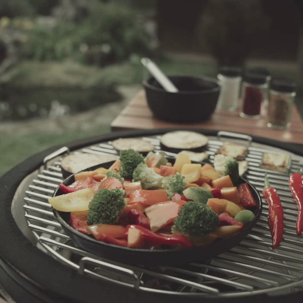 Vidéo présentant un poêlon en fonte noire pour kamado, filmé sous plusieurs angles. On y voit la fonte épaisse, la poignée amovible et son utilisation sur la grille du barbecue pour saisir, griller ou mijoter des aliments.