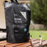 Sac de sciure de bois d’amandier posé sur une table en bois près d’un barbecue, accompagné d’une pince de cuisson en métal et bois au premier plan, dans un jardin.