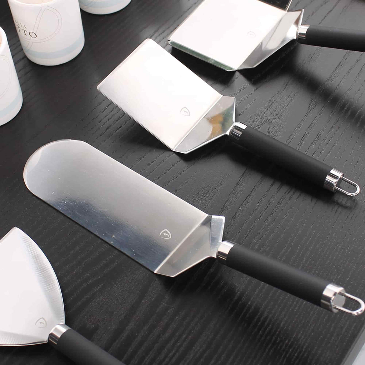 Ensemble de spatules en acier inoxydable avec manches noirs, disposées sur une table en bois foncé.