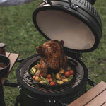 Poulet rôti placé verticalement sur un support de cuisson en fonte à l’intérieur d’un Kamado, entouré de légumes grillés colorés sur une grille au-dessus des braises.