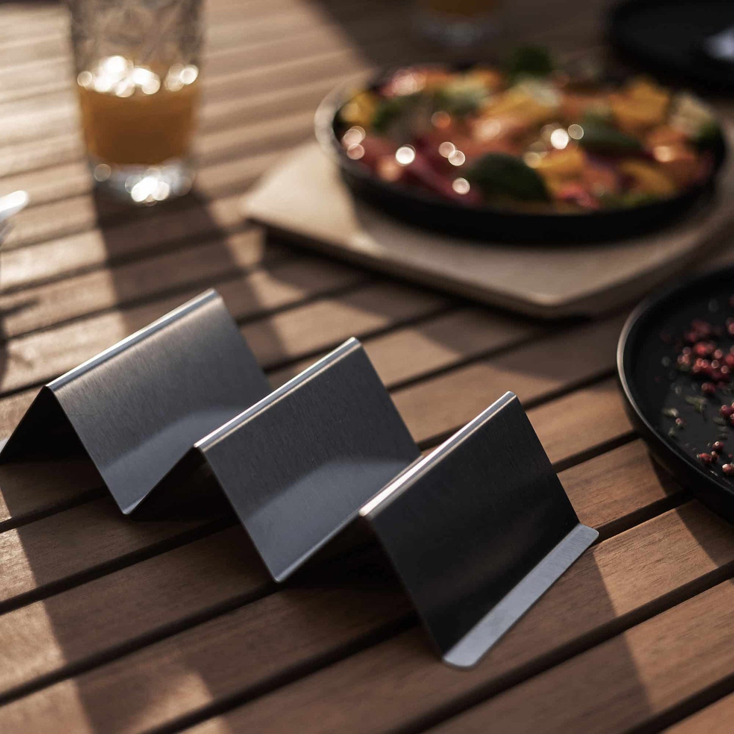 Support à tacos en inox brossé sur une table en bois, prêt à accueillir de délicieux tacos faits maison, avec des plats colorés en arrière-plan.