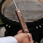 Main tenant un tire-cendre en acier inoxydable avec manche en bois gravé “Forest Grill”, devant un barbecue Kamado ouvert prêt à être nettoyé.