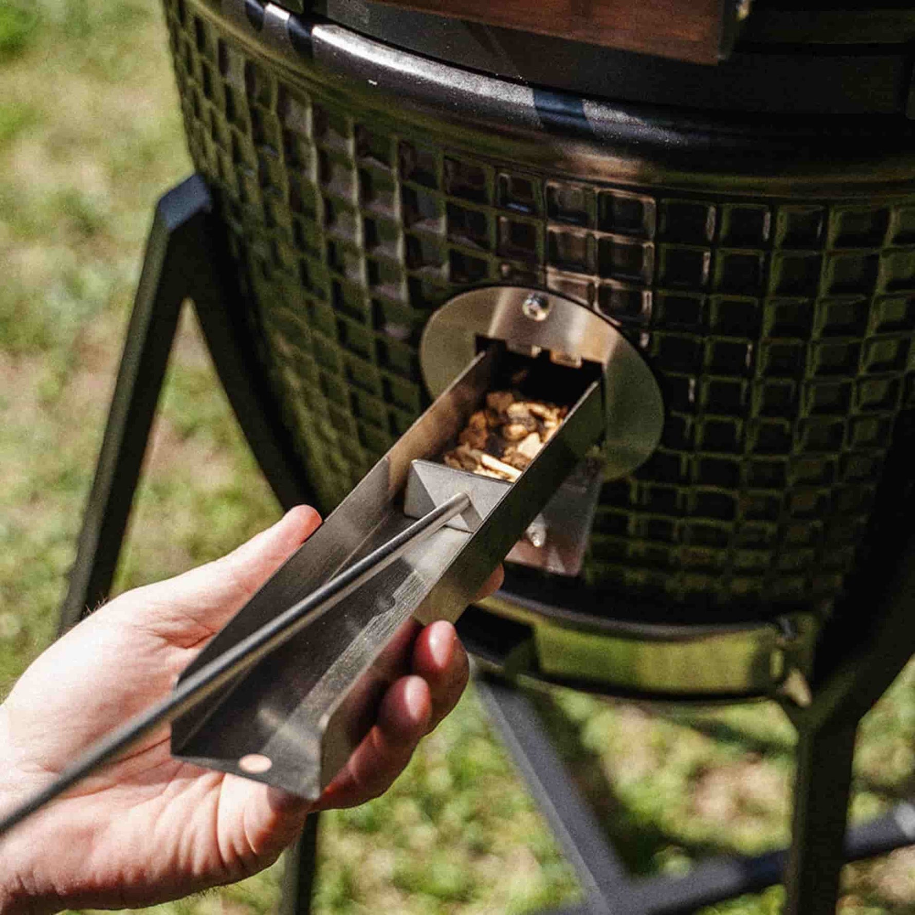 Main insérant un tiroir à copeaux de bois dans la trappe de fumage d’un barbecue Kamado, pour ajouter de la fumée sans ouvrir le couvercle.