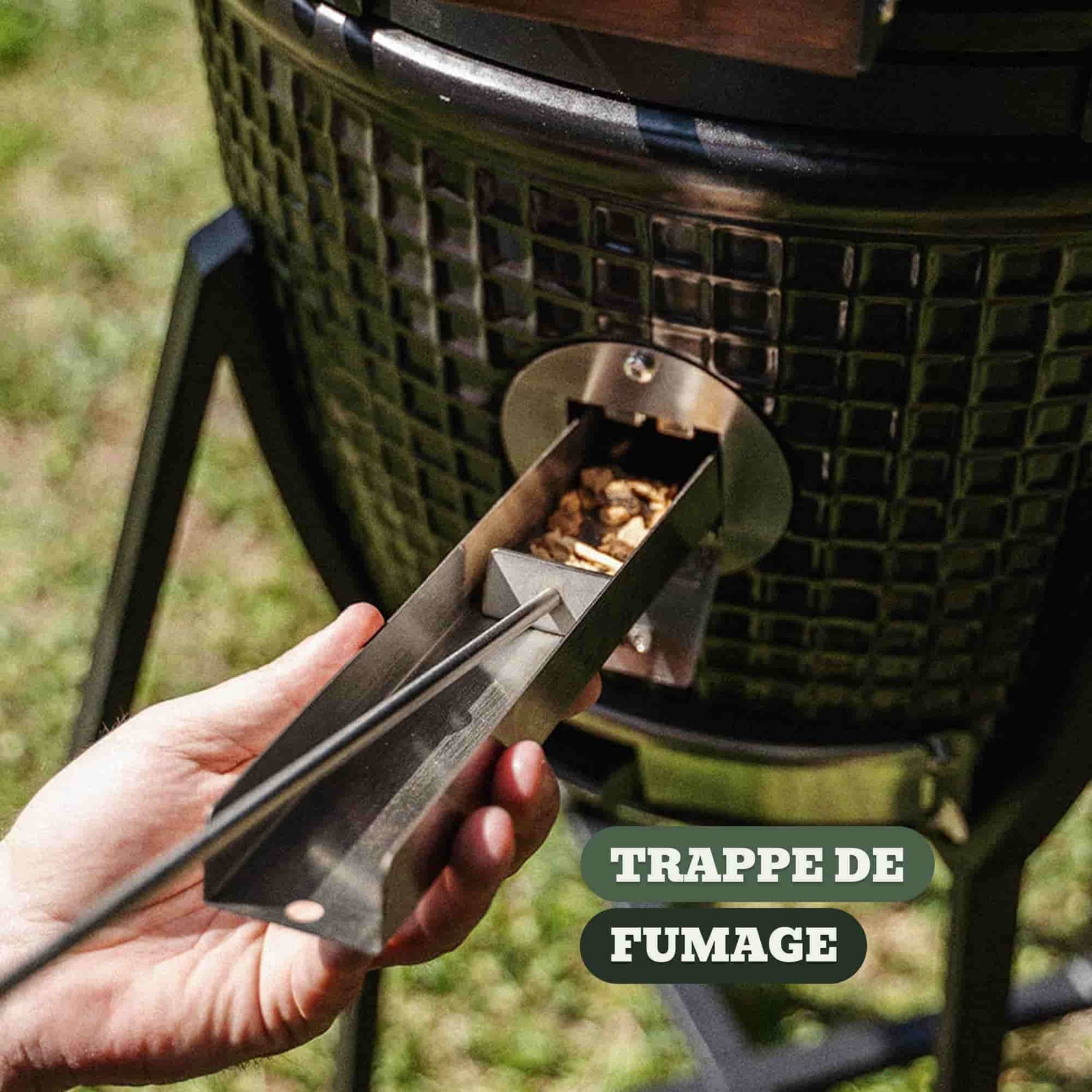 Main insérant des copeaux de bois dans la trappe de fumage en inox d’un barbecue Kamado, permettant d’ajouter de la fumée sans ouvrir le couvercle.