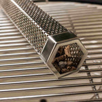 Tube générateur de fumée en acier inoxydable perforé, posé sur une grille de barbecue et rempli de copeaux de bois pour aromatiser les cuissons au Kamado.