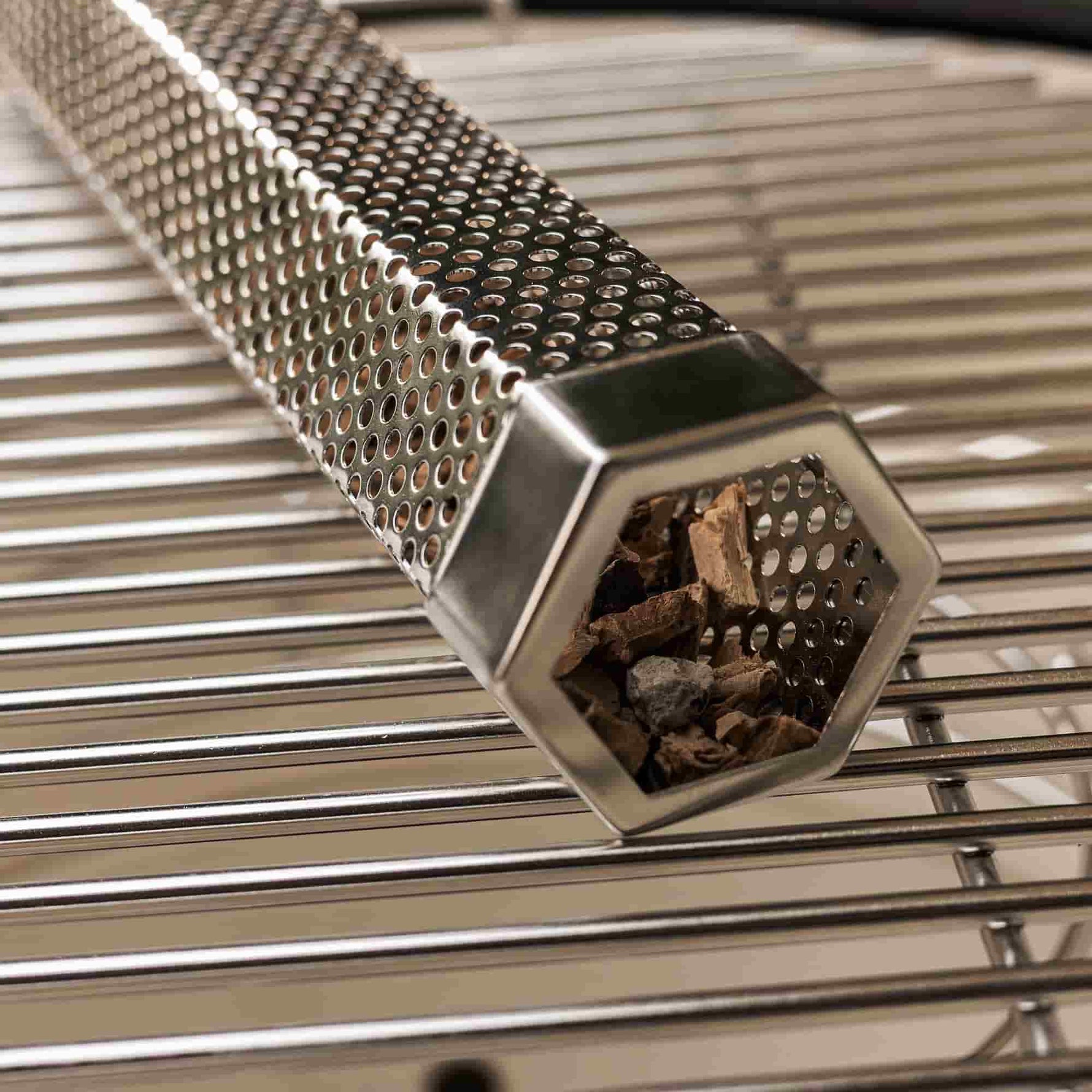 Tube générateur de fumée en acier inoxydable perforé, posé sur une grille de barbecue et rempli de copeaux de bois pour aromatiser les cuissons au Kamado.